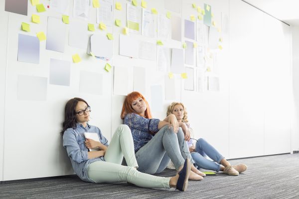 three women sitting against a wall with sticky notes and documents on wall