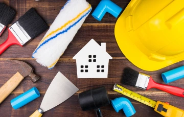 Tools like brushes, a hammer, and PVC pipes surround a white house cut-out on a wooden surface.