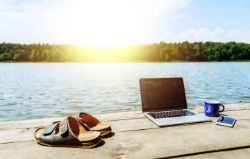 A laptop, sandals, coffee mug and a mobile phone laying a dock with the view of a lake