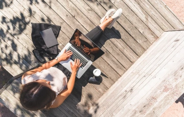 A woman sitting on a wooden deck outdoors working on a laptop