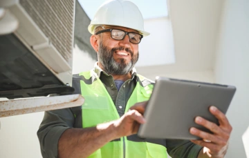 HVAC technician in uniform holding a tablet and smiling while standing in front of service equipment
