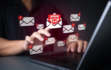 Illustration of a person’s hand interacting with a laptop as numerous email icons with red alert symbols float above it, with one envelope labeled “SPAM” featuring a skull—suggesting issues with email deliverability or performance.
