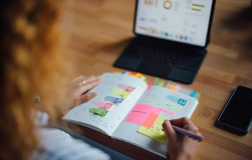 A person’s hands writing in a planner marked with multicolored sticky tabs, with a laptop showing charts and graphs visible in the background