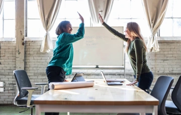 Two women high-five across a conference table with laptops and a rolled-up blueprint in a bright, industrial-style office, celebrating business success.