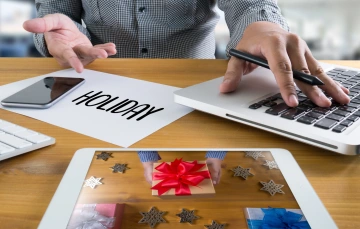 Hands working on a laptop and smartphone surrounded by holiday-themed items and a tablet displaying gift boxes