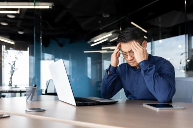 A man sitting in front of his laptop with his hands on his head and his eyes closed while looking frustrated.