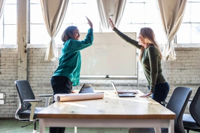Two women high-five across a conference table with laptops and a rolled-up blueprint in a bright, industrial-style office, celebrating business success.