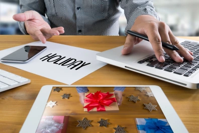Hands working on a laptop and smartphone surrounded by holiday-themed items and a tablet displaying gift boxes