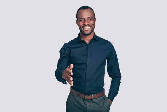 Handsome young African man stretching out hand for shaking and smiling to you while standing against grey background