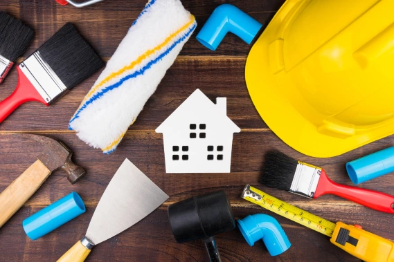 Tools like brushes, a hammer, and PVC pipes surround a white house cut-out on a wooden surface.