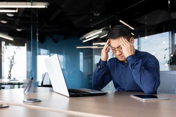 A man sitting in front of his laptop with his hands on his head and his eyes closed while looking frustrated.
