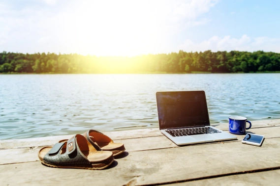 A laptop, sandals, coffee mug and a mobile phone laying a dock with the view of a lake