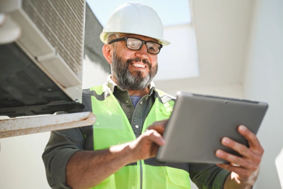 HVAC technician in uniform holding a tablet and smiling while standing in front of service equipment