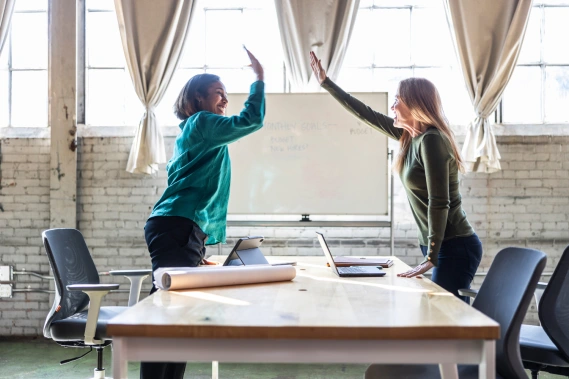 Two women high-five across a conference table with laptops and a rolled-up blueprint in a bright, industrial-style office, celebrating business success.